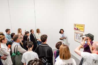 a woman stands in front of a Guerilla Girls work in front of a small crowd