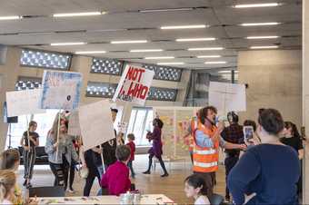 A group of people in a brightly lit museum space holding tall placards and walking around, with others at the side watching.