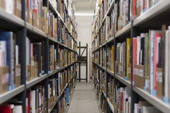 Two bookshelves containing cardboard file holders filled with numerous small books and pamphlets flank an aisle leading to an easel that has been propped up against the back wall