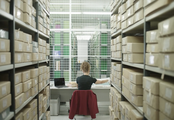 A person sitting at a desk with a laptop and books in a room stacked high with boxes on shelves